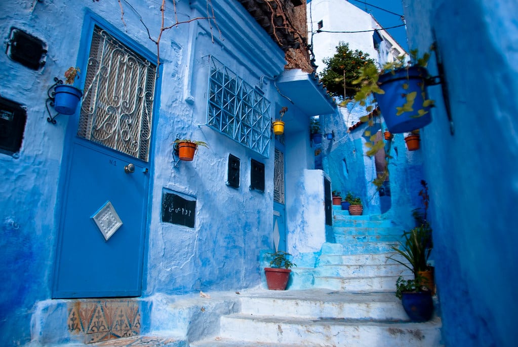 Um beco estreito com edifícios pintados de azul e plantas em vasos nas escadas em Chefchaouen, Marrocos, sob um céu azul claro.