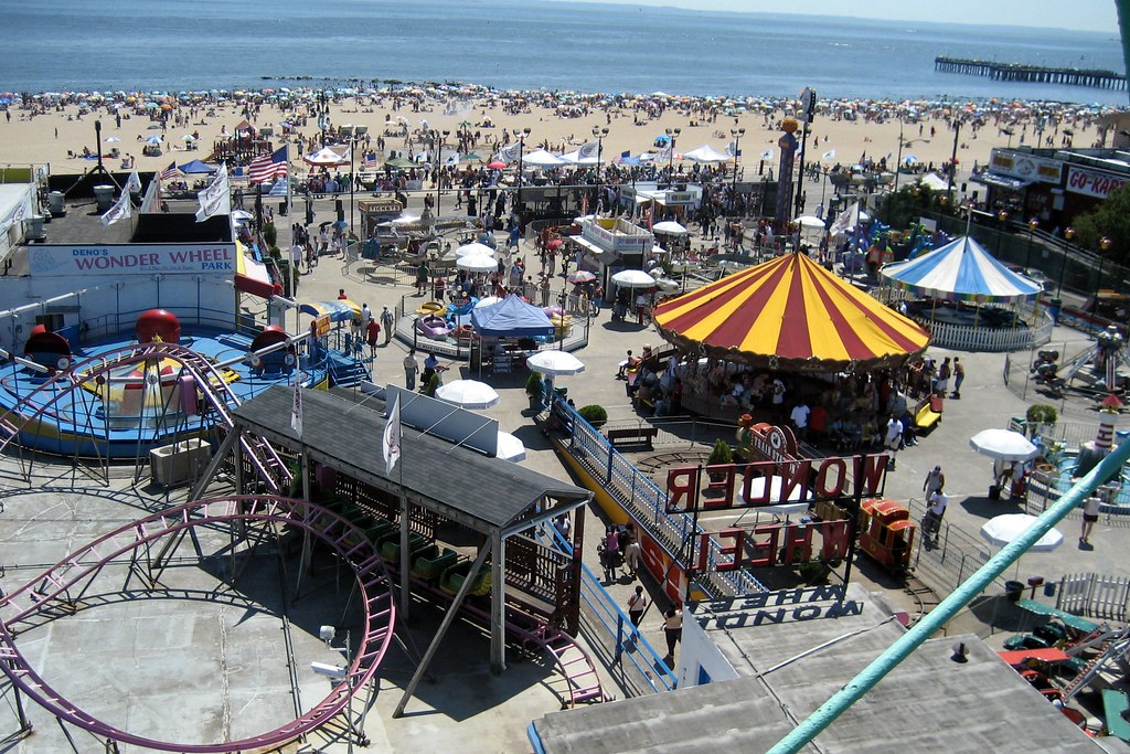 A imagem mostra uma cena movimentada de parque de diversões em Coney Island. Com uma roda-gigante, um carrossel e outros brinquedos, incluindo um com trilhos roxos. Há também uma grande multidão de pessoas na praia, aproveitando o clima ensolarado. Vários quiosques de comida e lanchonetes podem ser vistos, junto com calçadas cheias de visitantes. Foto para ilustrar post sobre o que fazer no Brooklyn.