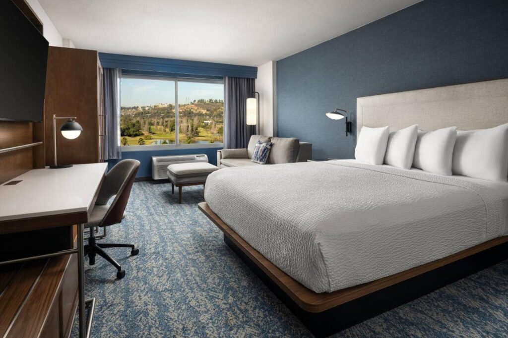 Quarto do Courtyard by Marriott. Uma cama de casal está do lado direito, de frente para uma TV e uma mesa de trabalho com cadeira. Ao fundo está um sofá e uma grande janela. A foto está no post de onde ficar em San Diego.