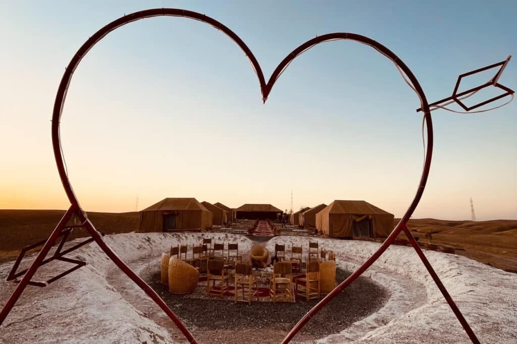 Escultura em forma de coração emoldura um acampamento no Deserto de Agafay com tendas marrons e uma área central com cadeiras e mesas ao pôr do sol.