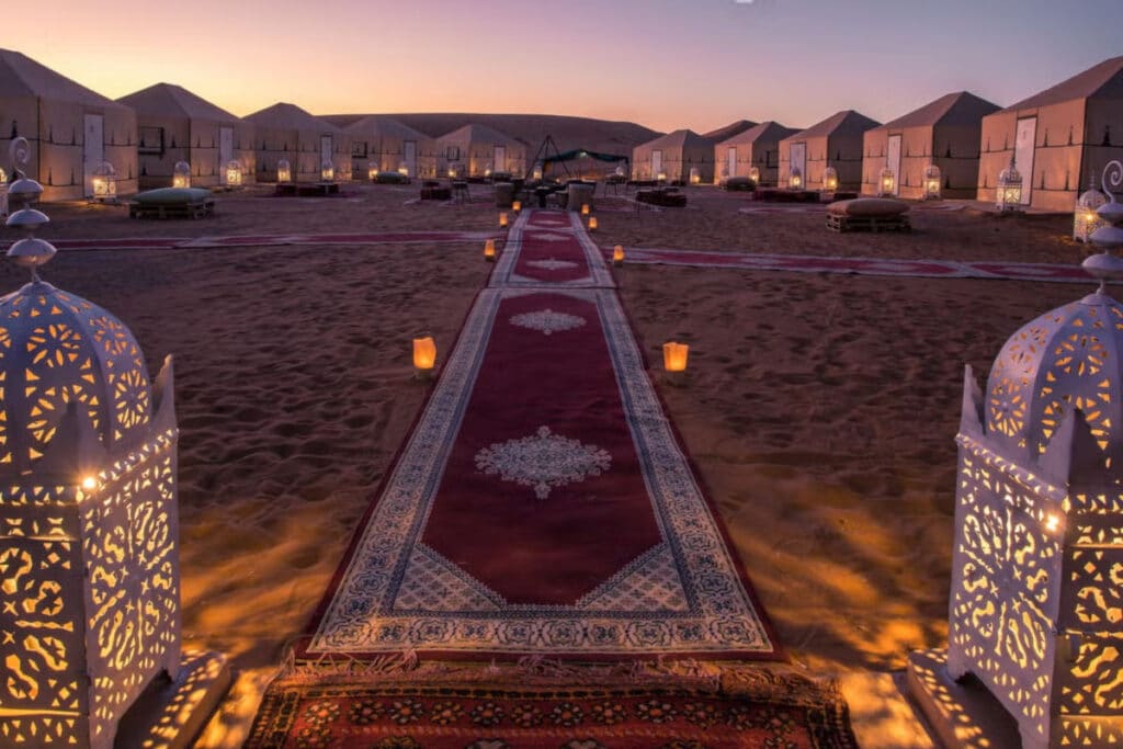 Acampamento elegante no deserto de Merzouga ao anoitecer com lanternas ornamentadas, um caminho acarpetado e tendas iluminadas ao fundo.