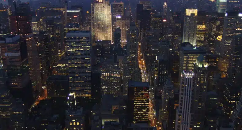 Vista aérea do Empire State Building de uma cidade à noite, com arranha-céus e ruas iluminados, estendendo-se à distância sob um céu escuro.