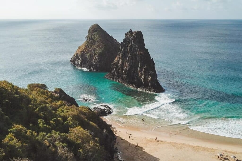 Vista aérea de uma da Enseada dos Abreus com formações rochosas no mar, água azul clara, folhagem verde e uma costa arenosa com pessoas. Está representando o post de melhores praias em Fernando de Noronha.