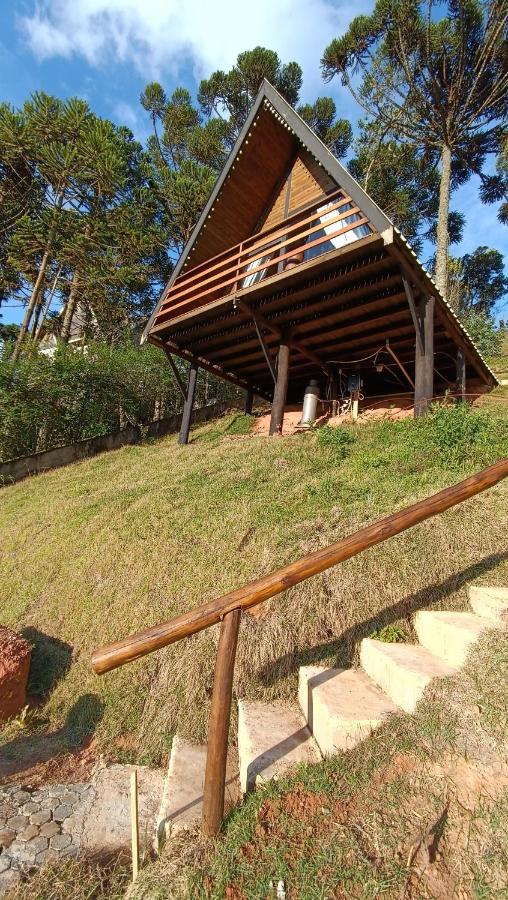 A imagem mostra uma cabana de madeira em formato triangular nos Chalés Capivari Diamante, situada em um terreno inclinado e cercada por árvores altas. A cabana está elevada sobre estacas, com uma varanda frontal protegida por um corrimão de madeira. Uma escada de pedra com um corrimão rústico leva até o nível da construção. O céu está claro, e o ambiente parece ser em uma área montanhosa ou campestre, rodeada por vegetação natural e grama. Esse local é uma recomendação entre os chalés em Campos do Jordão.