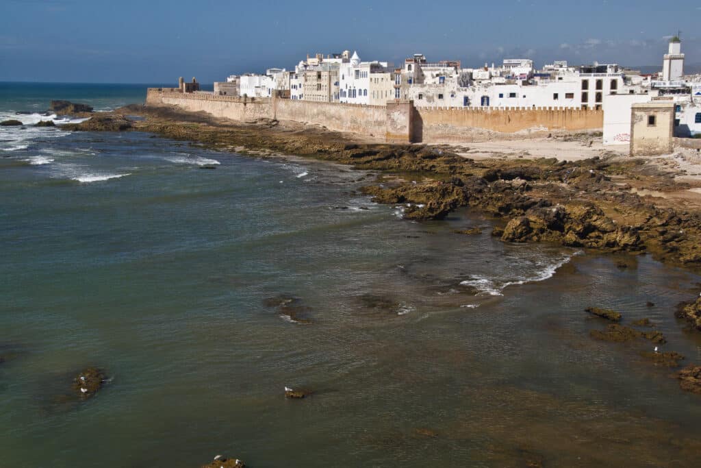 Vista costeira de Essaouira com edifícios brancos e uma muralha ao longo da costa rochosa, em direção ao oceano.