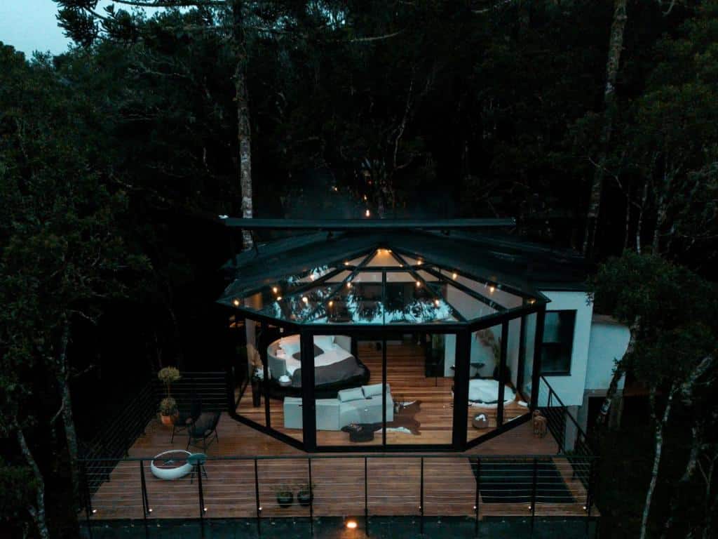 A imagem mostra uma vista aérea de um chalé do Benta Rosa Chalés de Luxo, localizado em uma área cercada por árvores, no meio de uma floresta. O chalé apresenta um design moderno e envidraçado, permitindo uma vista panorâmica do interior para a natureza ao redor. A estrutura possui um teto de vidro e paredes amplamente envidraçadas, criando uma sensação de integração com o ambiente natural. No interior, pode-se observar uma cama redonda e móveis sofisticados, oferecendo uma atmosfera de luxo e conforto. A varanda de madeira é espaçosa e conta com assentos ao ar livre, incluindo um pequeno espaço com uma banheira. O cenário ao redor é calmo, realçando a conexão com a natureza e o isolamento do local.