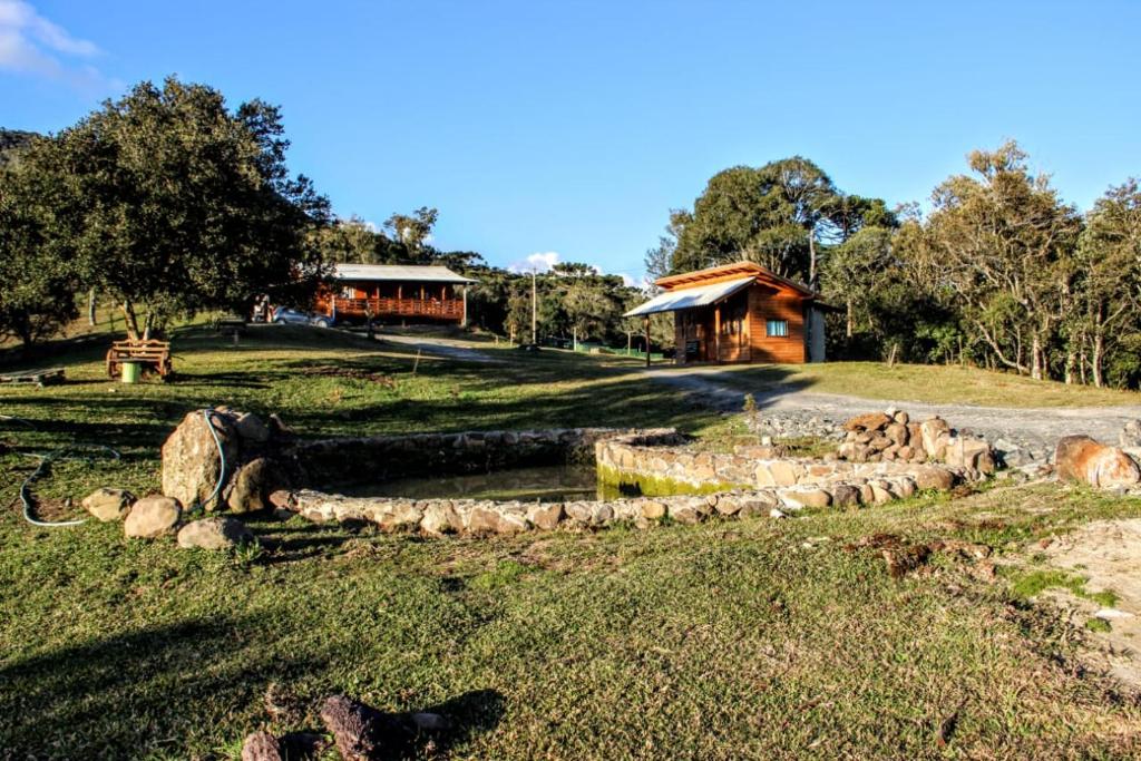 A imagem mostra o exterior da Pousada Recanto do Lobo Chalés em Urubici, localizada em uma área cercada por natureza, com árvores e vegetação ao redor. No terreno, há dois chalés de madeira, com telhados inclinados, situados em áreas abertas e espaçosas. Um pequeno lago artificial, rodeado por pedras, pode ser visto em primeiro plano. A paisagem transmite tranquilidade, com um amplo espaço ao ar livre ideal para descansar e apreciar a natureza.