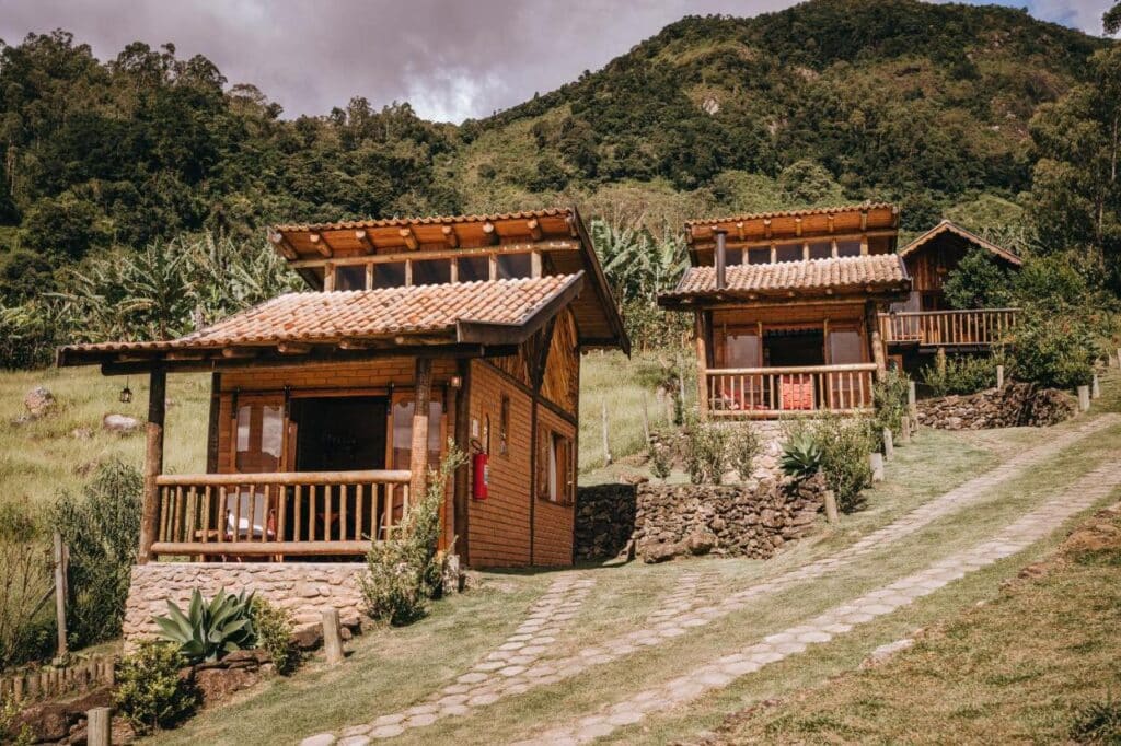 A imagem mostra dois chalés rústicos de madeira e pedra situados em uma encosta verdejante. Os chalés têm telhados de telha e varandas com cercas de madeira, com vista para um caminho de pedra que serpenteia pelo terreno inclinado. Ao fundo, há uma densa vegetação com árvores e colinas, criando uma atmosfera de tranquilidade em meio à natureza. O cenário é bucólico, com o verde das plantas contrastando com as construções de madeira clara, transmitindo uma sensação de isolamento e conforto. O Chalé Refugio da Pedra SP faz parte da lista de chalés para casal no interior de São Paulo.