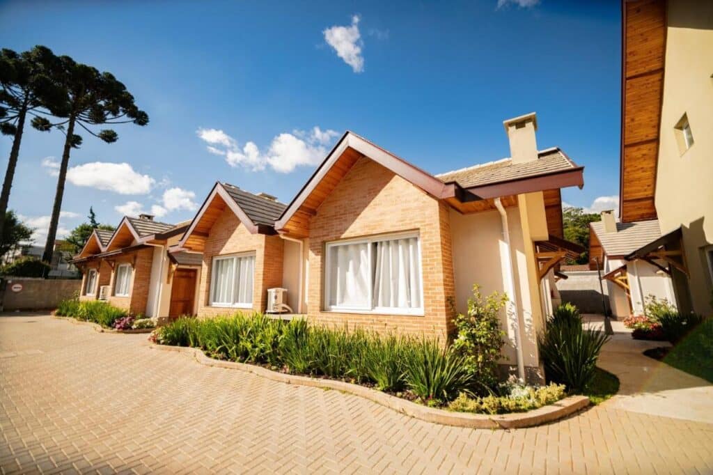 A imagem mostra uma fileira de casas geminadas nos Chalés Estilo de Campos, construídas com tijolos aparentes e telhados inclinados com acabamento de madeira. Cada casa possui uma grande janela com cortinas brancas e um pequeno jardim na frente, com plantas verdes bem cuidadas. O chão é pavimentado com blocos intertravados, formando uma calçada ampla e limpa. Ao fundo, há um céu azul claro com algumas nuvens dispersas, e ao lado esquerdo, altas árvores de pinheiro se destacam. A imagem transmite um ambiente organizado, ensolarado e acolhedor. O local faz parte da lista de chalés em Campos do Jordão.