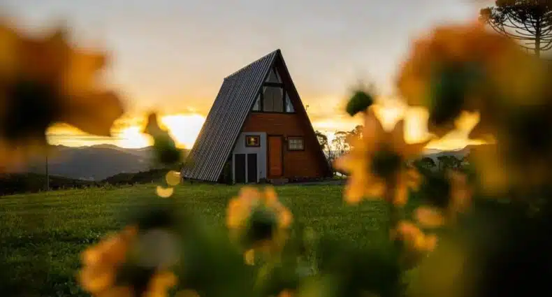 A imagem mostra uma cabana em formato triangular no Altitude 1140 Urubici - Chalés, um dos chalés em Urubici, localizado em um campo aberto, ao pôr do sol. A cabana é de madeira, com um teto inclinado que vai até o chão, típico de construções em forma de "A". Ao fundo, vê-se o céu dourado com o sol se pondo atrás das montanhas, criando uma atmosfera tranquila e acolhedora. Flores amarelas desfocadas em primeiro plano acrescentam um toque natural e harmonioso à cena.