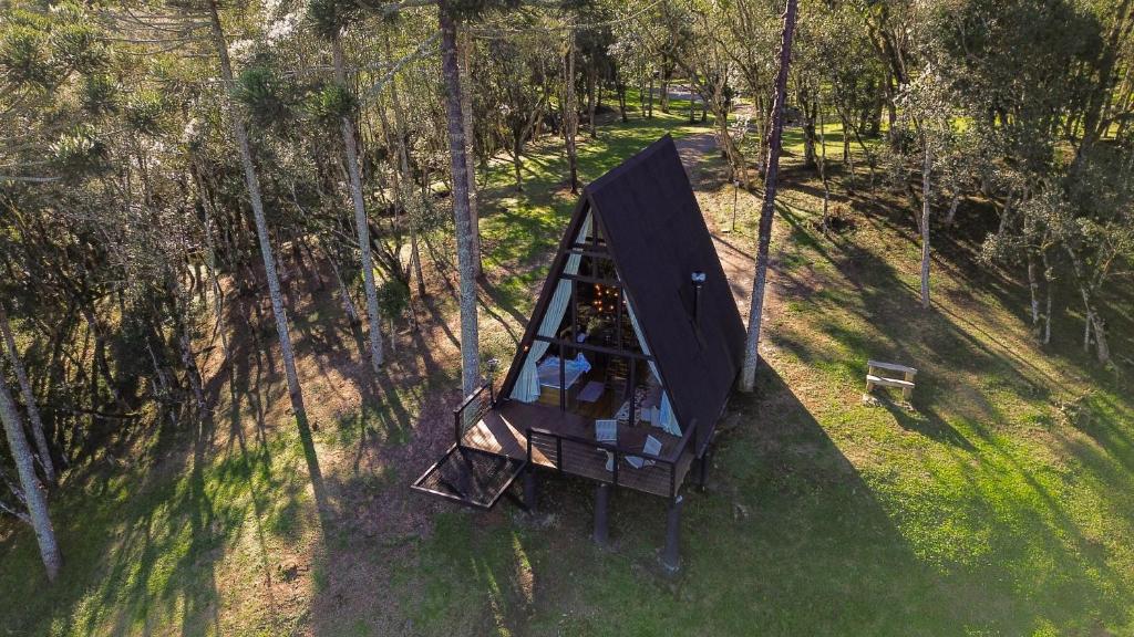 A imagem mostra uma vista aérea de um chalé no estilo "A-frame" (formato de A) localizado no meio de uma área arborizada no Hope Valley Chalés, em Urubici. O chalé é construído em madeira e está elevado em uma pequena plataforma com um deque na parte frontal. Ao redor, há uma vasta área verde com árvores altas, proporcionando um ambiente tranquilo e natural. Próximo ao chalé, pode-se ver um banco de madeira, sugerindo um espaço de relaxamento ao ar livre. O design do chalé é simples e integrado à paisagem natural ao redor.