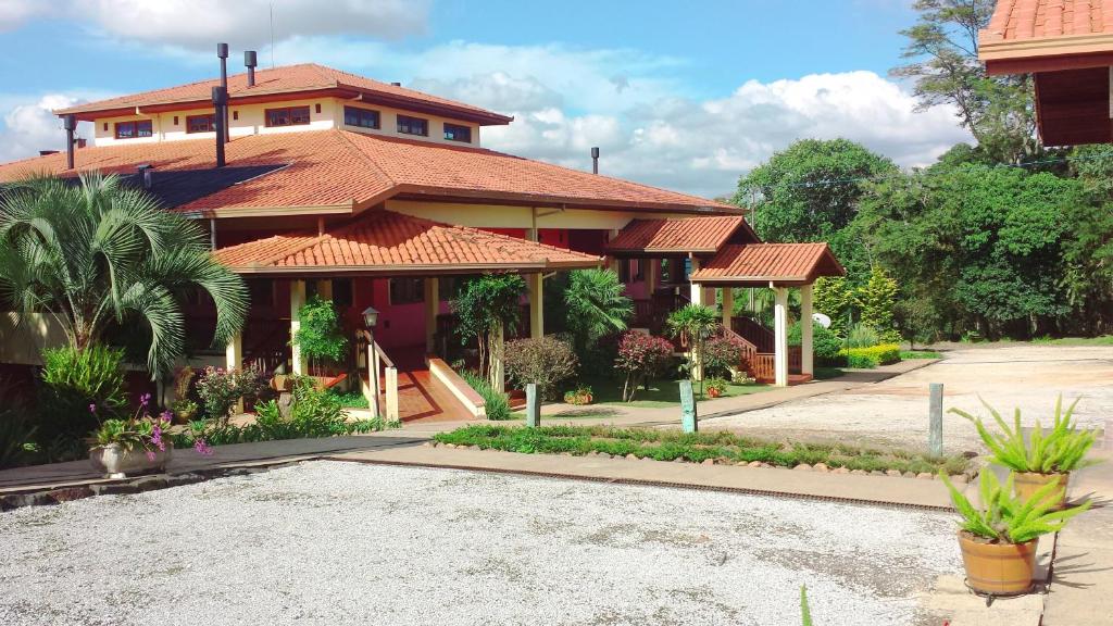 A imagem mostra a fachada do Hotel Fazenda Itáytyba, no Paraná, uma construção ampla e charmosa com telhado de telhas vermelhas. O edifício tem um design acolhedor, com varandas cobertas e pilares que sustentam a estrutura. O jardim ao redor é bem cuidado, com vegetação variada, incluindo palmeiras e plantas ornamentais. O céu azul claro e algumas nuvens ao fundo completam a paisagem, enquanto a vegetação ao redor e as árvores indicam a presença de áreas verdes e tranquilidade, características de uma fazenda.