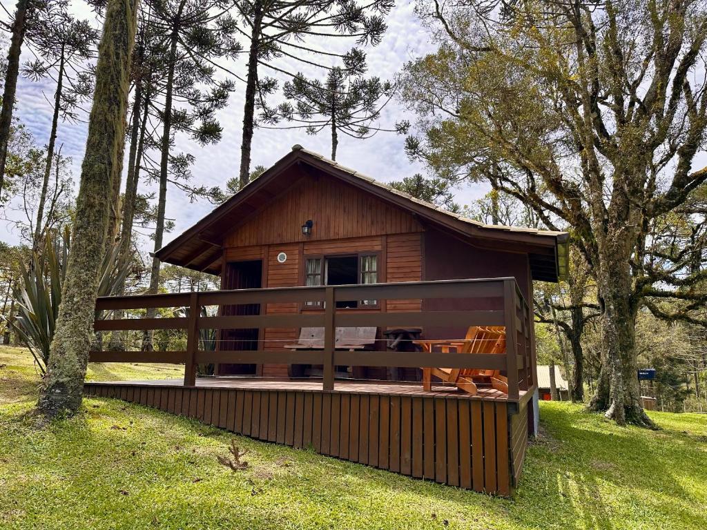 A imagem mostra um chalé de madeira em Urubici, o Namastê - Hospedaria de Altitude, em meio a uma área arborizada com araucárias. O chalé possui uma varanda com cerca de madeira, equipada com cadeiras de descanso. A construção de estilo rústico combina com o ambiente natural ao redor, e o gramado ao redor é bem cuidado. O local parece ser tranquilo, cercado pela natureza.