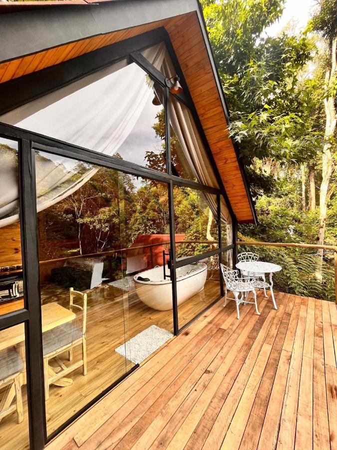 A imagem mostra a área externa do chalé Platanus com grandes janelas de vidro que permitem ver o interior. Do lado de fora, há um amplo deck de madeira com uma mesa e cadeiras brancas de ferro, criando um espaço confortável para relaxar ao ar livre. Dentro, há uma banheira branca próxima à janela, oferecendo uma vista para a vegetação ao redor. O ambiente está cercado por árvores e natureza, proporcionando uma sensação de tranquilidade e conexão com o ambiente natural. A decoração mistura elementos rústicos e modernos. Esse local é uma recomendação entre os chalés em Campos do Jordão.