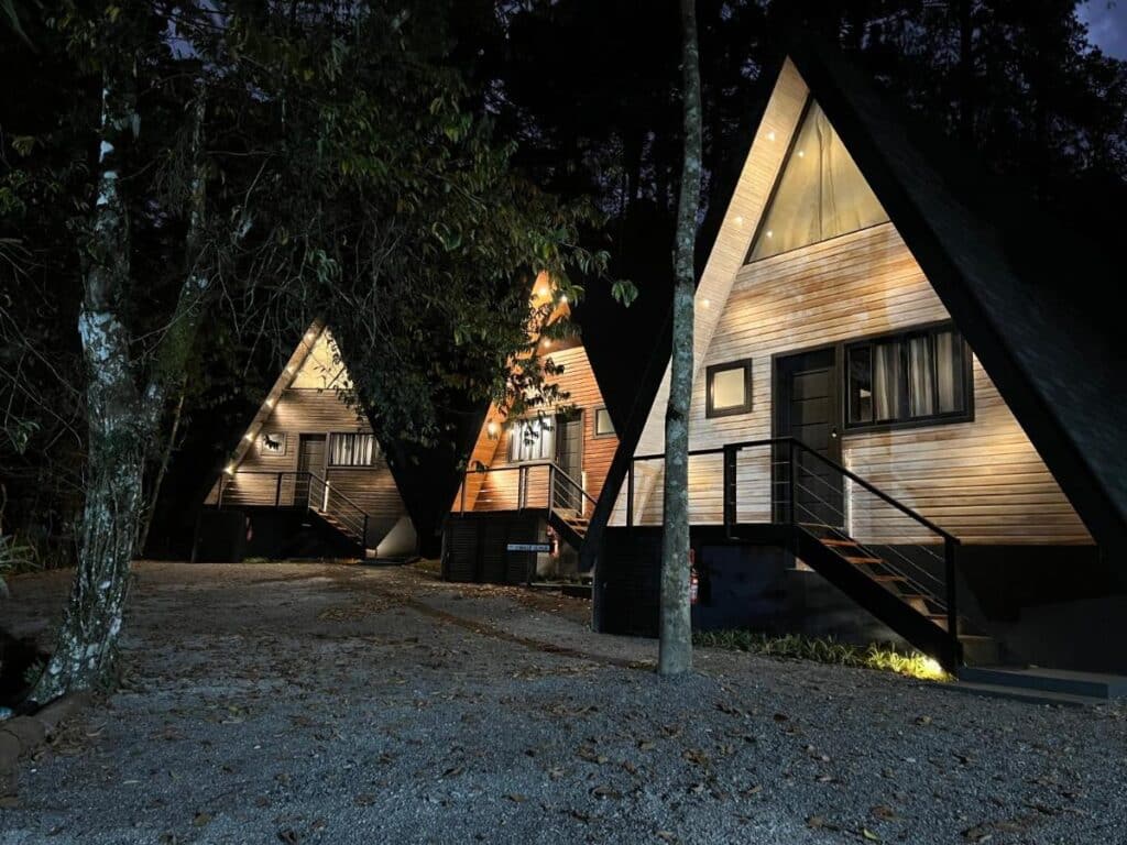 A imagem mostra três chalés em formato de "A", iluminados durante a noite em meio a uma área arborizada no Chalé Villa Nature. As cabanas têm um design moderno com paredes de madeira clara e janelas retangulares. Cada chalé possui uma pequena escada que leva à porta de entrada, com corrimões pretos de metal. A iluminação suave destaca o formato triangular das construções, criando uma atmosfera acolhedora e tranquila. A área ao redor está coberta por folhas secas e árvores, reforçando o ambiente natural e isolado.