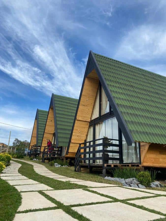 A imagem mostra uma fileira de cabanas em formato triangular nos Chalés entre Nuvens, com telhados verdes e paredes de madeira clara, situadas em um gramado bem cuidado. As cabanas possuem grandes janelas de vidro na parte frontal e pequenas varandas de madeira com corrimão preto. Um caminho de pedras brancas com grama entre os blocos leva até as cabanas. O céu está claro com algumas nuvens finas, criando um ambiente tranquilo e natural. Uma pessoa de casaco vermelho está de pé em uma das varandas, observando a paisagem ao redor. Esse local é uma recomendação entre os chalés em Campos do Jordão.