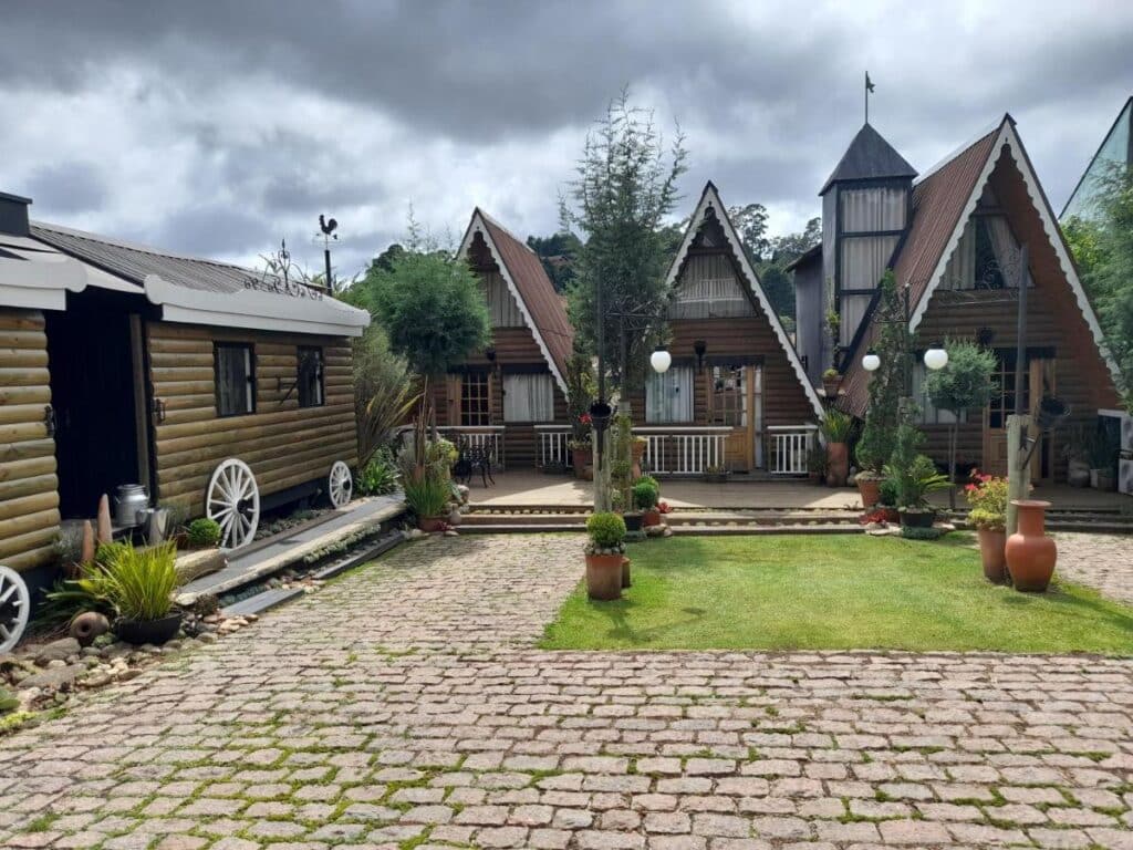 A imagem mostra um pátio de uma propriedade com chalés de madeira em estilo rústico nas Suítes no Chalé. Três chalés triangulares com telhados inclinados estão ao fundo, com janelas grandes e detalhes decorativos. À esquerda, há uma construção com formato de carroça antiga, com rodas brancas e paredes de madeira. O chão é de pedras irregulares, cercado por vasos de plantas e pequenos jardins bem cuidados. O céu está nublado, criando uma atmosfera serena, e há várias árvores e vegetação ao redor, complementando o ambiente campestre e acolhedor. Esse local é uma recomendação entre os chalés em Campos do Jordão.