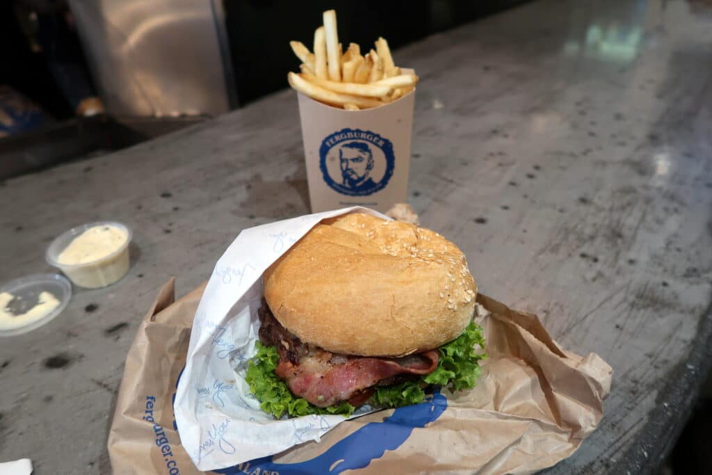 Um hambúrguer do Fergburger, em Queenstown, com alface e bacon em uma embalagem. Está ao lado de uma xícara de batatas fritas em um balcão cinza.
