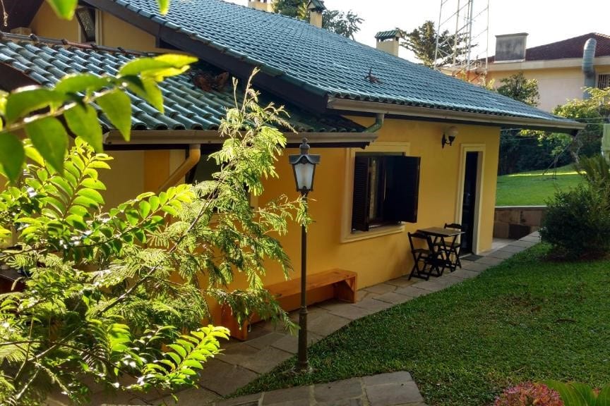 Vista da frente do Chalé no Centro de Canela durante o dia; as parede em tom amarelo com porta e janelas de madeira e em volta há grama com arbustos. Representa chalés em Canela.