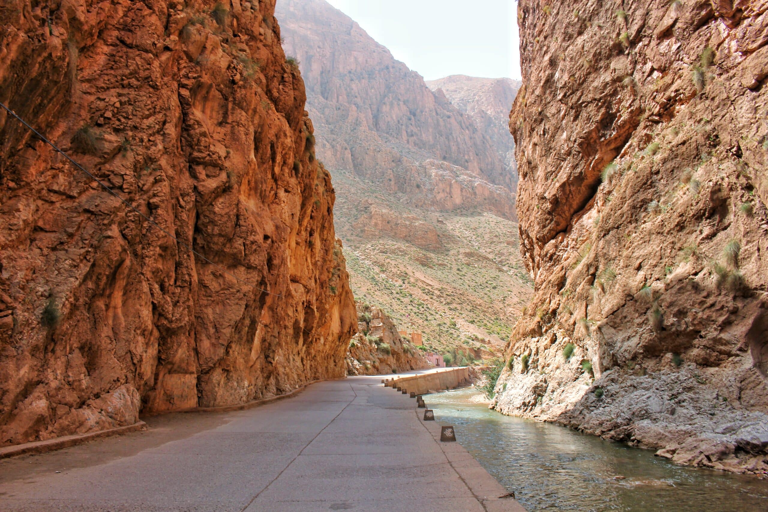 Uma estrada estreita passa entre imponentes penhascos rochosos com um pequeno rio correndo ao lado sob um céu claro em Gorges du Todra.
