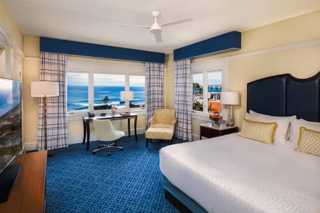 Quarto do Grande Colonial La Jolla com uma cama de casal do lado direito, em frente a uma TV. Ao fundo estão uma mesa com cadeira, uma poltrona e uma janela com vista para o mar. Esse hotel é outra boa opção de onde ficar em San Diego.