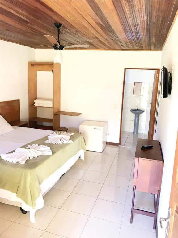 Quarto do Hotel Fazenda Bandeirantes. Uma cômoda e uma TV do lado direito, no fundo a porta do banheiro. Do lado esquerdo uma cama de casal, no fundo um frigobar e uma estante. No teto um ventilador.