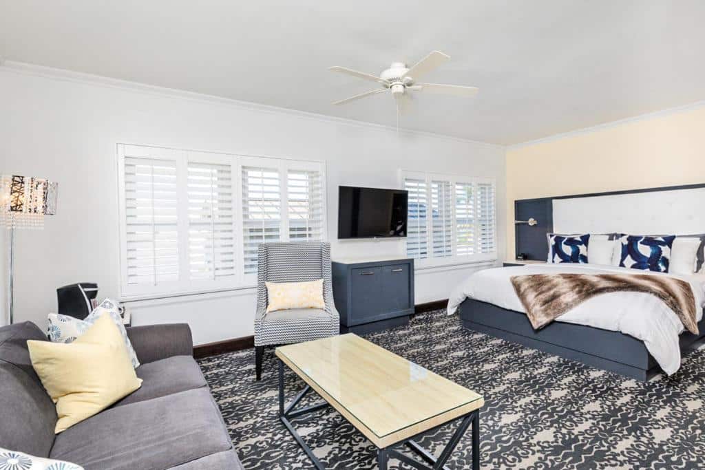 Quarto do Hotel Marisol com uma cama de casal do lado direito, de frente para um sofá, uma poltrona e uma mesa de centro. Ao fundo estão duas janelas e uma TV.