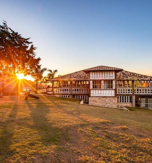 A imagem mostra um grande casarão de estilo colonial em uma paisagem montanhosa ao pôr do sol. O edifício tem janelas amplas e paredes brancas com detalhes em pedra, além de um telhado de telhas tradicionais. À esquerda, o sol se põe atrás de uma árvore, lançando longas sombras sobre o gramado ao redor. O céu está claro, com tons dourados e laranjas. Ao fundo, é possível ver as montanhas e o horizonte, criando uma vista panorâmica deslumbrante. O ambiente transmite tranquilidade e beleza natural. Esse é o Hotel Vila Relicário, um dos hotéis-fazenda em Minas Gerais.