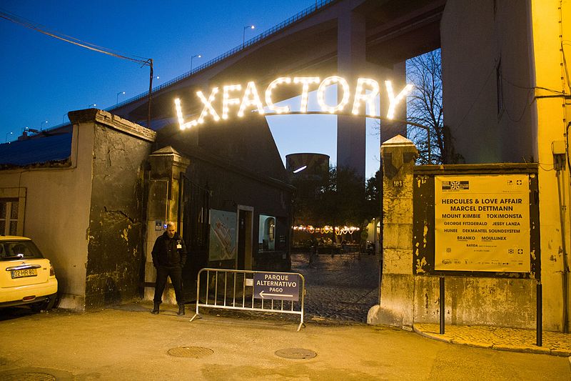 Imagem da frente da entrada do LX Factory á noite com um segurança do lado direito na porta de entrada.