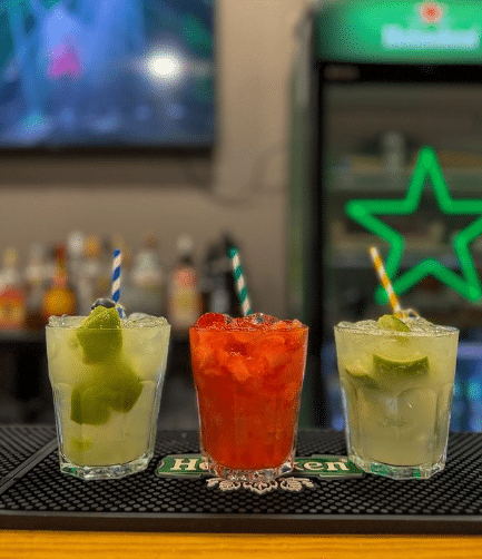 Três bebidas com canudos listrados no Maria Bonita em um balcão de bar, duas com limão e uma vermelha no meio, garrafas de bebidas e geladeira ao fundo.