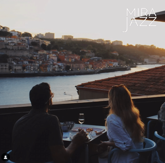 Duas pessoas jantando em um restaurante na cobertura com vista para o rio e a cidade ao pôr do sol. O texto "MIRA JAZZ" está no canto superior direito.