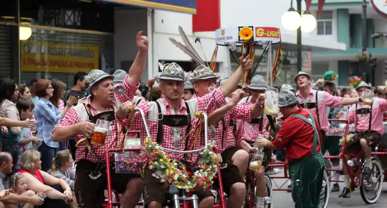 Um grupo de homens vestindo lederhosen andam de bicicleta com vários assentos, segurando canecas de cerveja no Oktoberfest Blumenau. Imagem para ilustrar post sobre o que fazer em Blumenau.