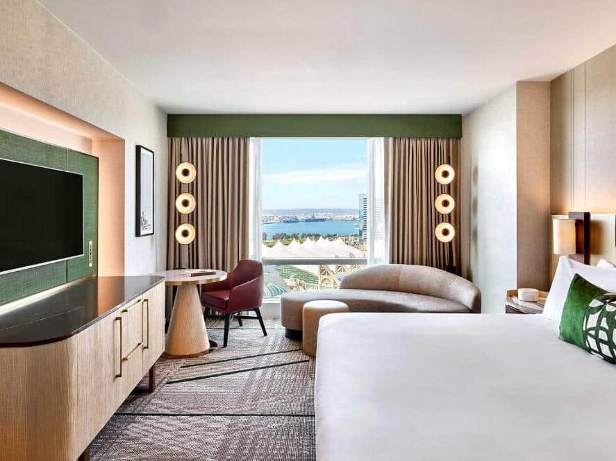 Quarto do Omni San Diego Hotel. Uma cama de casal está do lado direito. Em frente está um armário com balcão e uma TV. Ao fundo está uma grande varanda e um sofá. A foto está no post de onde ficar em San Diego.