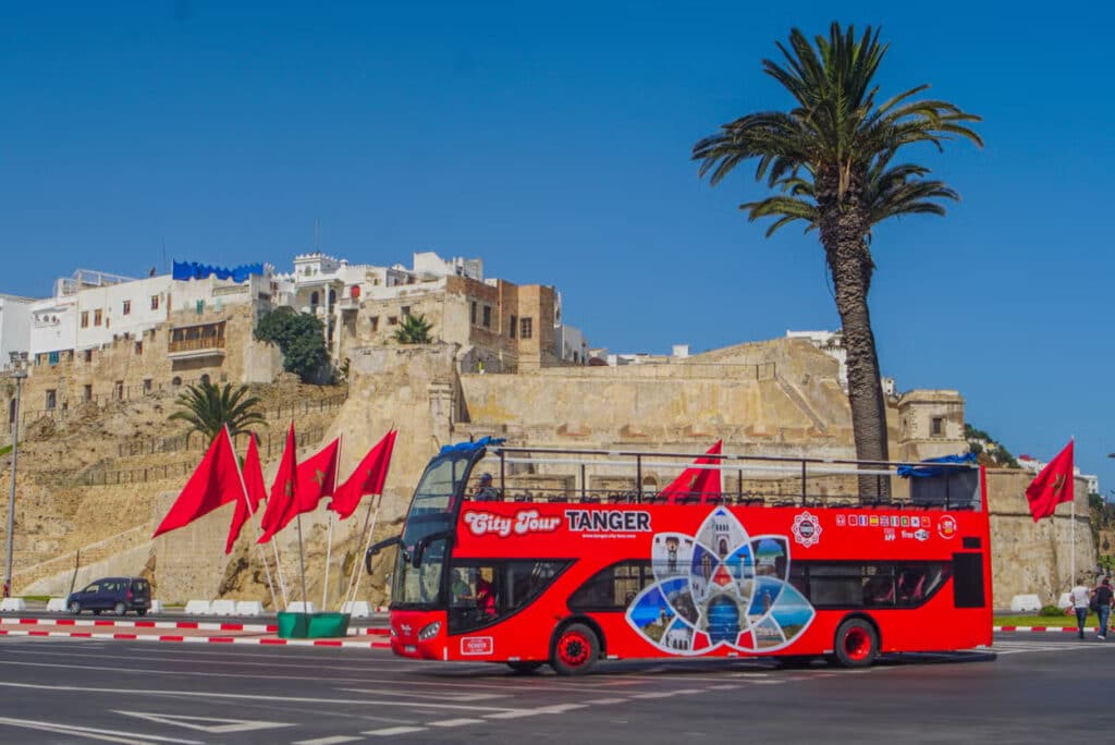 Um ônibus turístico vermelho em Tânger de dois andares passa por um edifício histórico com várias bandeiras vermelhas e palmeiras ao redor. Imagem para ilustrar post sobre o que fazer no Marrocos.