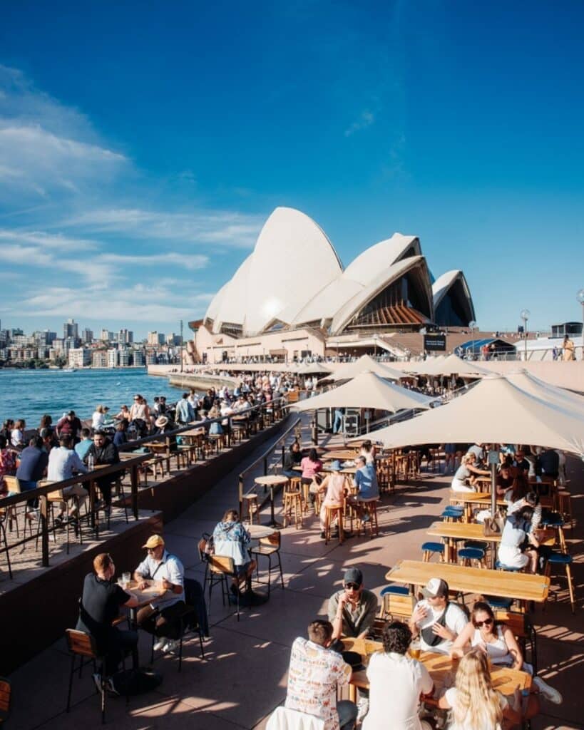 Pessoas jantam ao ar livre em mesas iluminadas pelo sol perto da Sydney Opera House. O céu está limpo, e a paisagem urbana é visível ao fundo. Está no Opera Bar em Sydney.