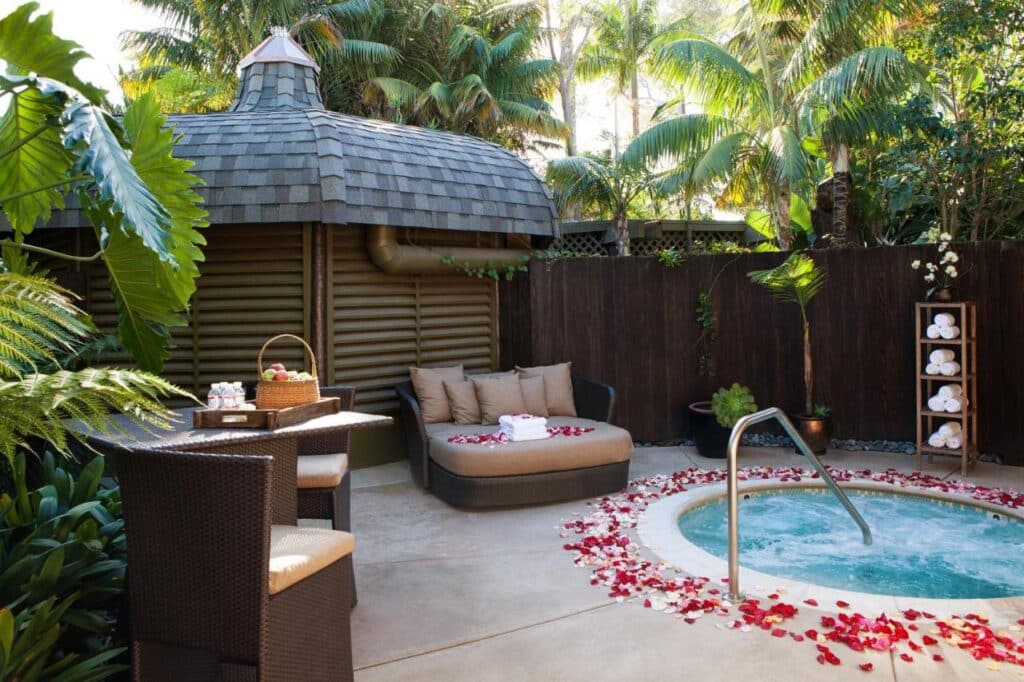 Área de spa do Paradise Point. Uma banheira redonda, ao ar livre, está no canto direito de frente para um sofá. Várias petalas de flor estao espalhadas ao redor. Ao fundo está um lugar fechado. Na frente dele há uma mesa com cadeiras.