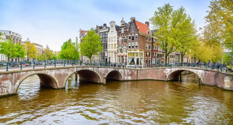 Uma ponte de pedra se ergue sobre um canal ladeado por edifícios tradicionais e árvores sob um céu azul em Amsterdam. Imagem para ilustrar post sobre o que fazer em Amsterdam.