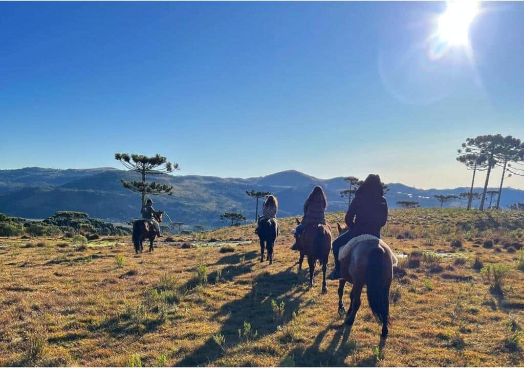 Imagem de quatro pessoas andando de cavalo a frente do Fazenda Santa Rita Turismo Rural durante o dia com montanhas ao fundo.