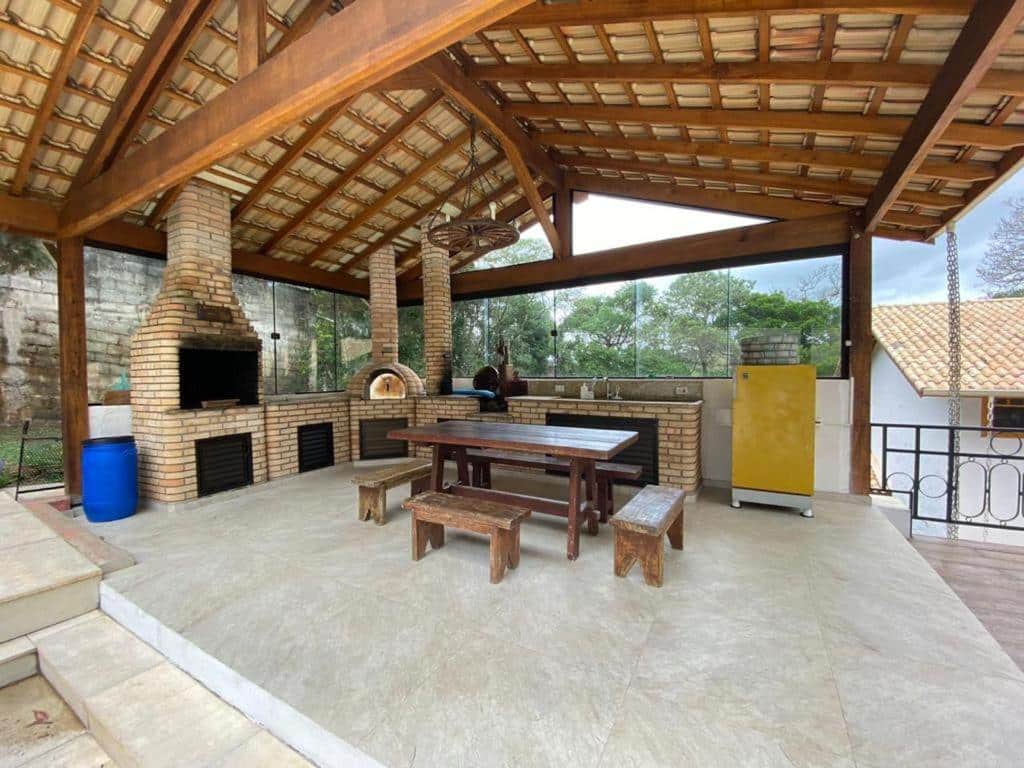 Varanda do Paz e Aconchego. Uma mesa de madeira no meio, do lado direito uma geladeira, no fundo uma pia e um forno de pizza e do lado esquerdo uma churrasqueira.