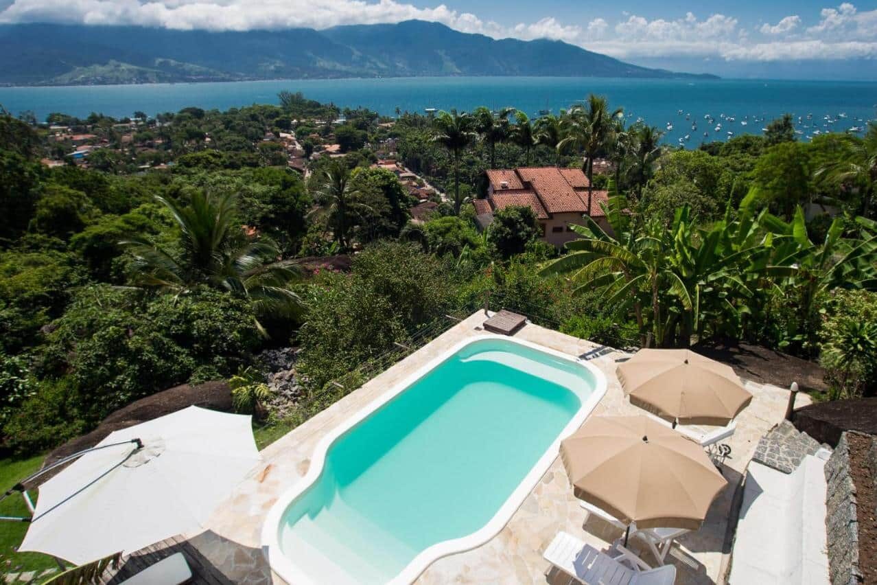 A imagem mostra uma piscina ao ar livre com água clara, cercada por cadeiras de descanso e guarda-sóis nos Chalés Mariolinha. A piscina está localizada em uma área elevada, proporcionando uma vista panorâmica de uma paisagem tropical com árvores verdes densas, telhados de casas ao longe, e o mar ao fundo. Além do mar, há montanhas cobertas por vegetação e parcialmente encobertas por nuvens. O cenário transmite uma sensação de tranquilidade e beleza natural, com céu azul e tempo ensolarado.
