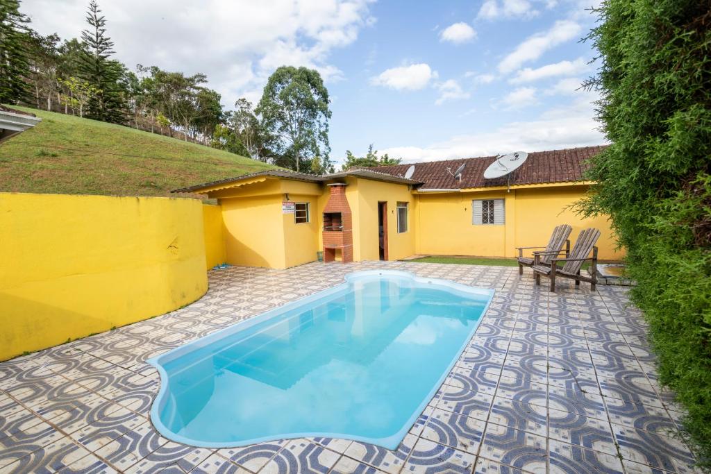A imagem mostra uma área externa com uma piscina de tamanho médio cercada por um piso de azulejos com padrões geométricos em azul e branco nos Chalés Tia Nastácia. As paredes ao redor são pintadas de amarelo, criando um ambiente vibrante. Ao fundo, há uma pequena construção também amarela, com uma churrasqueira de tijolos à vista. Duas cadeiras de madeira estão posicionadas ao lado da piscina para descanso. O cenário é rodeado por uma colina verde e algumas árvores, enquanto o céu está parcialmente nublado, proporcionando uma atmosfera tranquila.