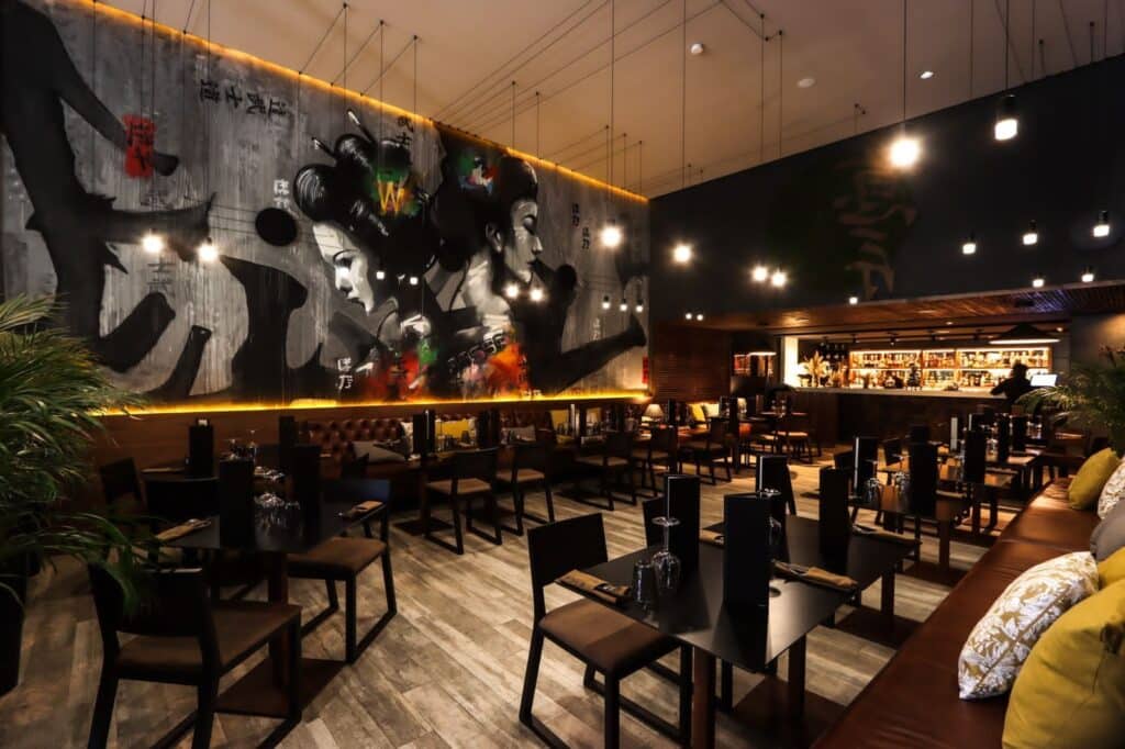 O restaurante Poka com pouca iluminação e decoração moderna, com um mural de gueixas na parede e mesas com cardápios e copos. Imagem para ilustrar post sobre o que fazer no Marrocos.