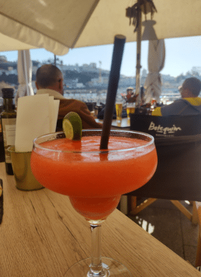 Uma bebida vermelha congelada brilhante do Botequim Nostalgic decorada com uma rodela de limão e um canudo preto repousa sobre uma mesa de madeira em um café ao ar livre com pessoas sentadas. Representa o que fazer à noite no Porto.