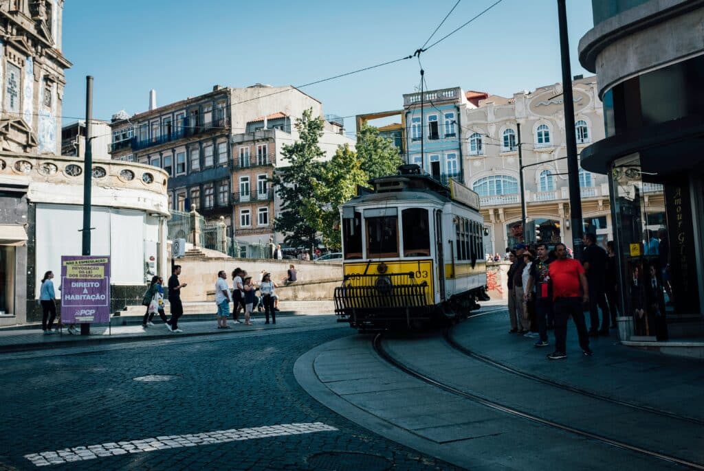 Um bonde amarelo e branco circula por uma esquina no Porto em uma área urbana, com vários pedestres por perto em um dia claro e ensolarado. Representa o que fazer à noite no Porto.