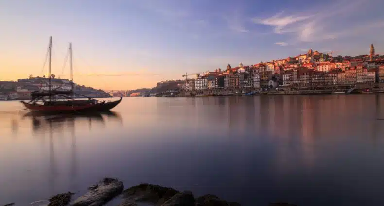Imagem do rio Douro no Cais da Ribeira, no final do dia com o rio do lado esquerdo e um barco navegando e do lado direito ao fundo o Cais da Ribeira. Representa onde comer na cidade do Porto.