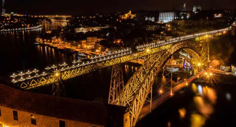 Uma grande ponte em arco iluminada atravessa um rio à noite, com inúmeras luzes e edifícios visíveis em ambos os lados da água. Representa o que fazer a noite no Porto.