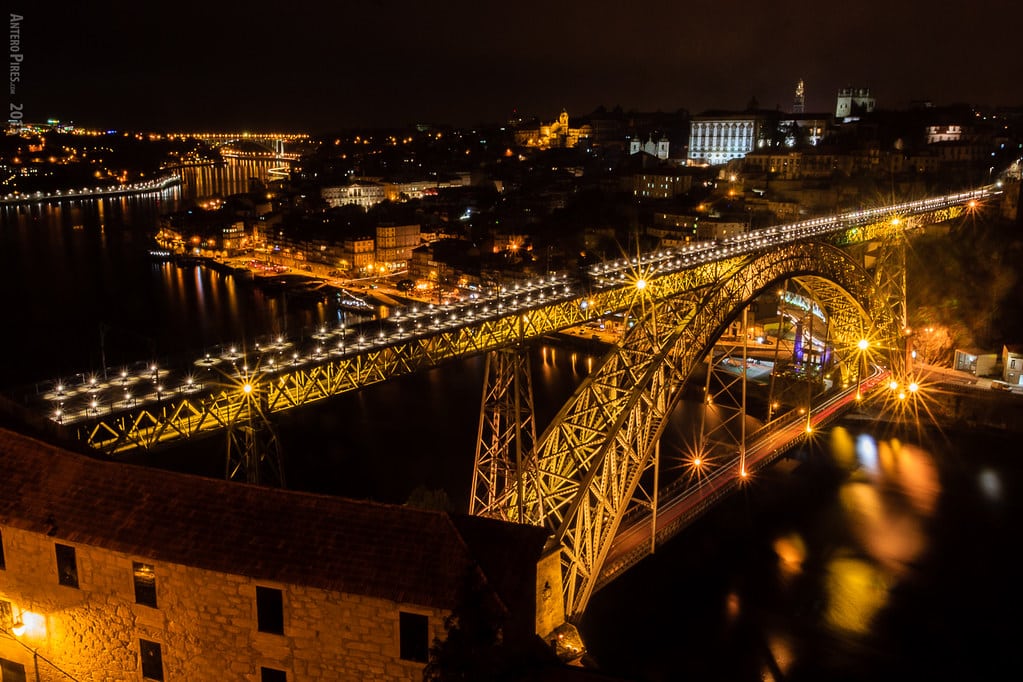 Uma grande ponte em arco iluminada em Porto atravessa um rio à noite, com inúmeras luzes e edifícios visíveis em ambos os lados da água. É a Ponte D. Luis que representa um dos roteiros para Portugal.