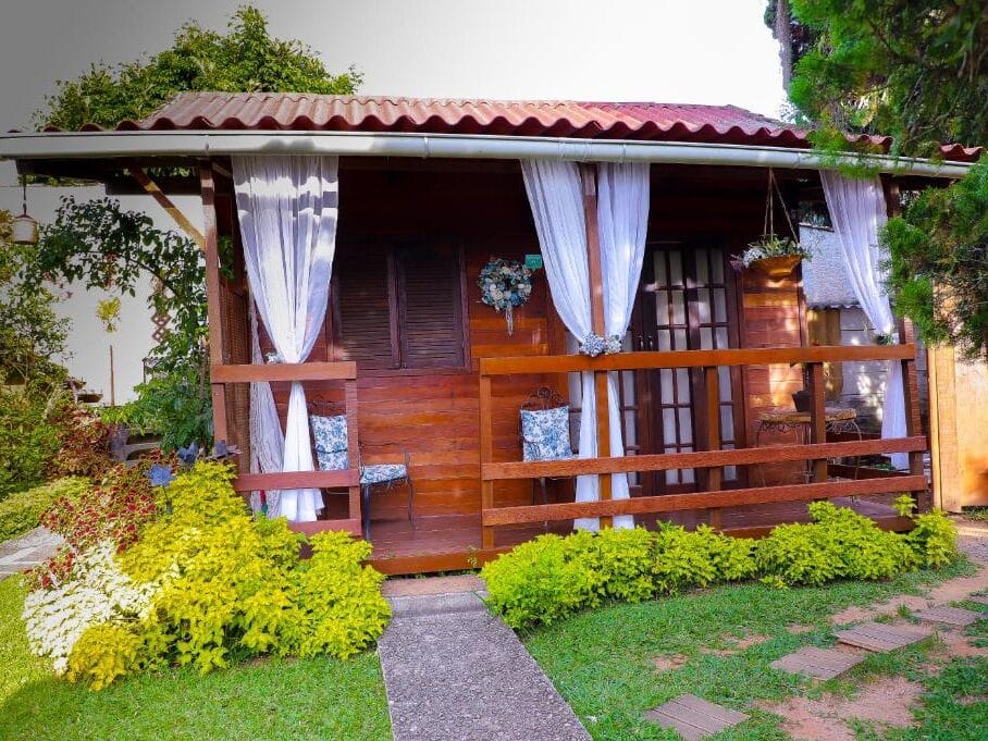 Lado de fora de um chalé na Pousada do Lago. É uma pequena cabana de madeira com cortinas brancas, uma varanda, vasos de plantas e um caminho de jardim verdejante que leva à entrada.