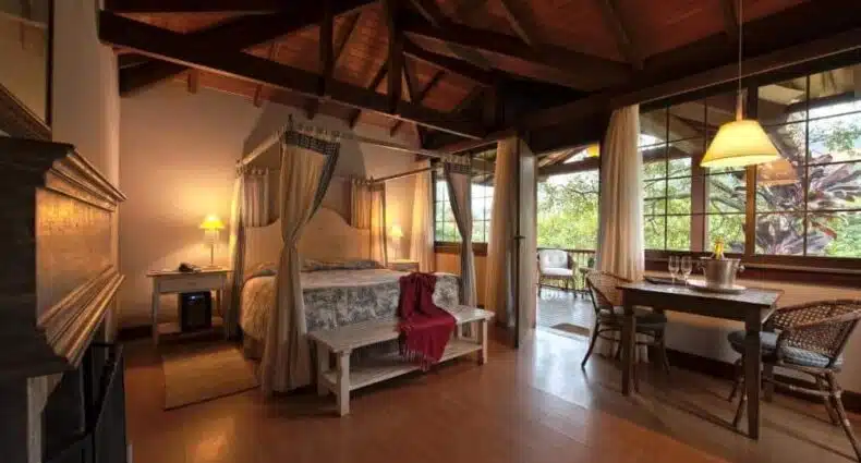 Quarto da Pousada Les Roches. Uma cama de casal está no meio, perto de uma mesa de jantar. Ao seu lado direito está uma grande parede de vidro e porta que leva até a varanda. O teto é de madeira. A imagem ilustra o post de chalés em Petrópolis.