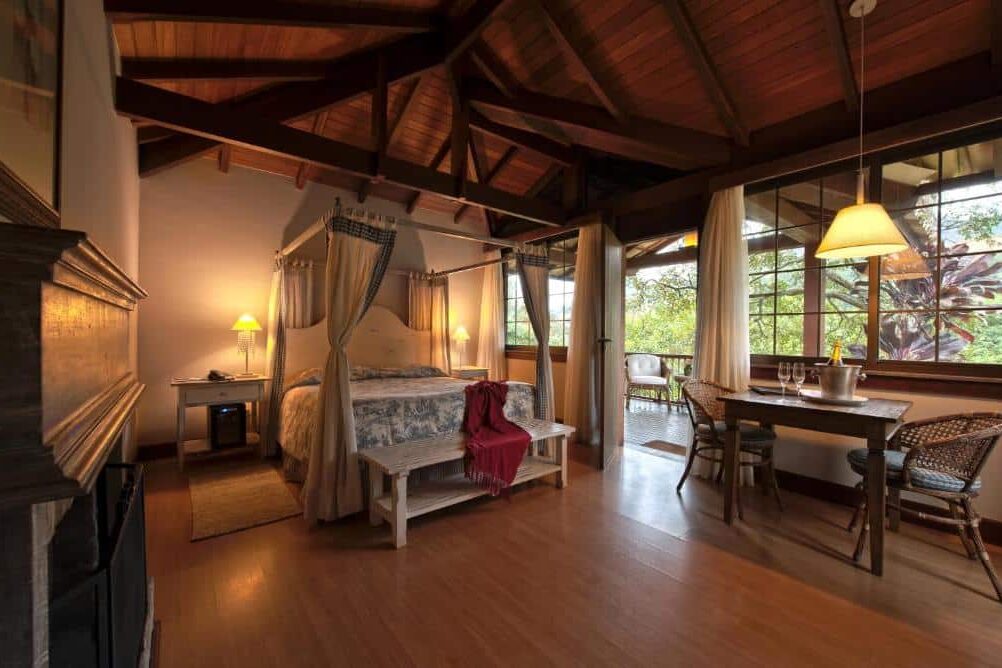 Quarto da Pousada Les Roches. Uma cama de casal está no meio, perto de uma mesa de jantar. Ao seu lado direito está uma grande parede de vidro e porta que leva até a varanda. O teto é de madeira. A imagem ilustra o post de chalés em Petrópolis.